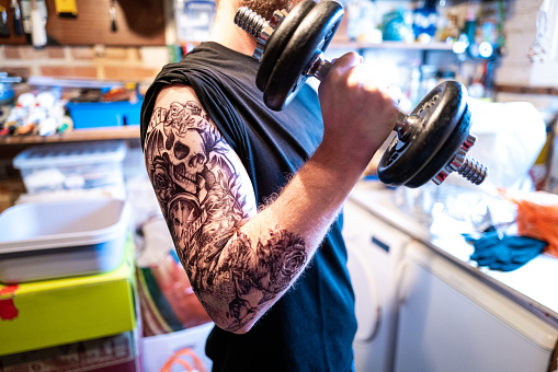 Color image depicting a mid adult man in his 30s lifting weights in his garage. The man has a sleeve tattoo on his arm. Room for copy space.