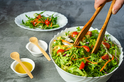 Man's hand serving salad of lettuce, tomatoes, rugula, various green vegetables with wooden tongs to an oval dish. White bowl full of salad bowl full of salad and small plates with salad dressing.