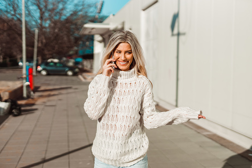 Smiling young woman with long blond hair wearing a white sweater talking on a smartphone in the city street on a sunny winter day