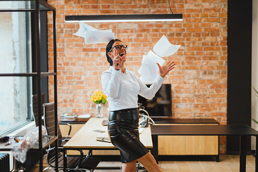 Happy and smiling fashionable businesswoman throwing paperwork in the air, celebrating success and working in the modern office with a red brick wall