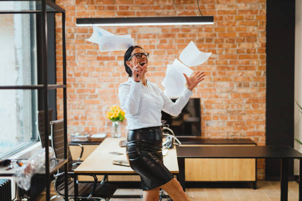 Happy businesswoman throwing papers in the air in the office Happy and smiling fashionable businesswoman throwing paperwork in the air, celebrating success and working in the modern office with a red brick wall quitting a job stock pictures, royalty-free photos & images