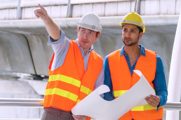 istock Two engineer or architect with safety helmet planning about building with blueprint. Professional technician man pointing finger while his colleagues looking at construction site.Industry work concept 1302617304