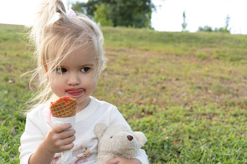 Cute little caucasian child licking ice cream, holding bear toy, sitting on green grass, summer time, tasty ice cream, happy childhood concept.
