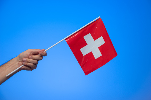Small Swiss national flag held by Caucasian male hand isolated on blue background studio shot.