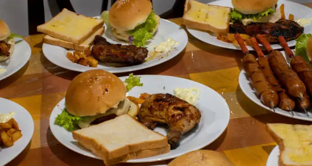 Photo of Dinner plates ready at the kitchen table, Family sits together for dinner time. serving bbq chicken with burgers, and grilled sausages with bread.