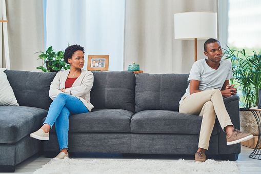 Shot of a young couple sitting apart on a couch after an argument while she looks at him in disdain