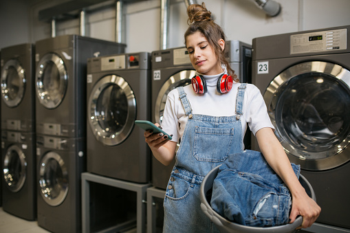 Attractive young Caucasian woman doing her laundry at the laundromat place, using a smart phone to entertain herself while her laundry's being washed
