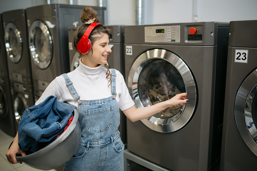 Attractive young Caucasian woman doing her laundry at the laundromat place, having her headphones on and listening to music while her laundry's being washed
