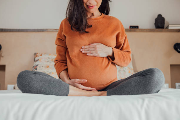 Cropped Pregnant woman, sitting in crossed legs in be at home holding her belly. Front view of cropped long hair brunet caucasian pregnant woman, sitting in crossed legs, in yoga pose, in bed at home, holding her belly with hand. Maternity, pregnancy, new life. pregnant stock pictures, royalty-free photos & images