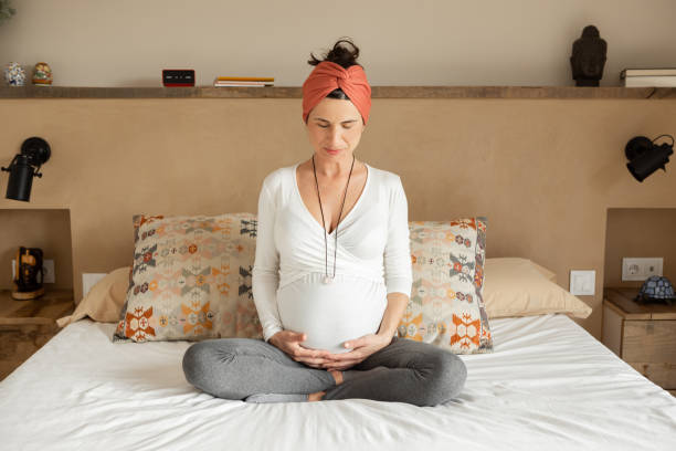 mulher grávida relaxada sentada em pose de ioga na cama com os olhos fechados - human pregnancy pensive women thinking - fotografias e filmes do acervo