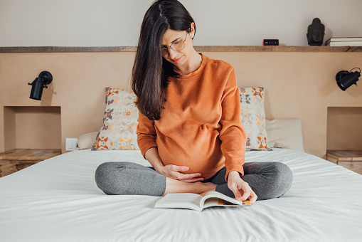 Front view of Beautiful long hair brunet caucasian pregnant woman, wearing glasses, sitting in crossed legs in bed, reading a book and holding her belly with hand. Maternity, pregnancy, new life.