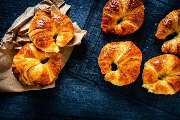 Overhead view of six fresh homemade croissants shot on dark blue background. Four croissants are on a cooling rack and two placed directly on the table. Predominant colors are yellow and blue. High resolution 42Mp studio digital capture taken with SONY A7rII and Zeiss Batis 40mm F2.0 CF lens