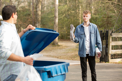 Mid-adult Latin Descent and young Caucasian men put recycle trash in bin.  The young adult brings out a few more items to add to the recycle collection.  Bin is located on curb in front of home and is picked up twice a week.