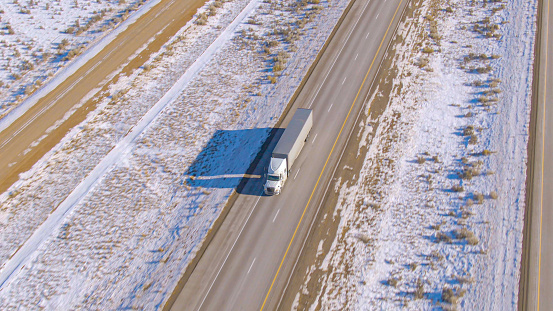DRONE: White truck hauls cargo down a scenic asphalt freeway running across the snowy desert in Utah. Snowy desert surrounds the highway leading a big rig across the rugged United States wilderness.