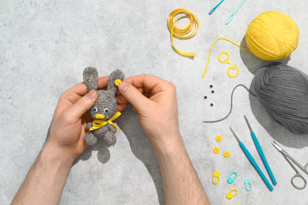 istock DIY Easter gift making theme. Human hands hold handmade knitted toy Easter rabbit and needlework accessories on grey background. Knitting concept. Overhead view, flat lay 1302662495