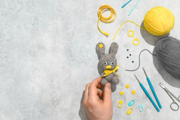 istock DIY Easter gift making theme. Male hand hold handmade knitted toy Easter rabbit and needlework accessories on grey background. Knitting concept. Overhead view, flat lay, copy space 1302662501