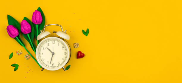 istock Spring background with decorative flowers and alarm clock, yellow flat lay banner, copy space 1302669438