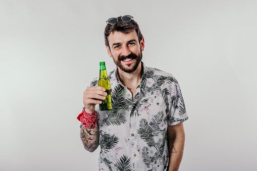 Portrait of a caucasian man wearing a Hawaiian style floral shirt, holding a beer bottle, and looking at the camera. A young guy in a summer-style outfit against a gray background. Tropical holiday and alcohol concept.