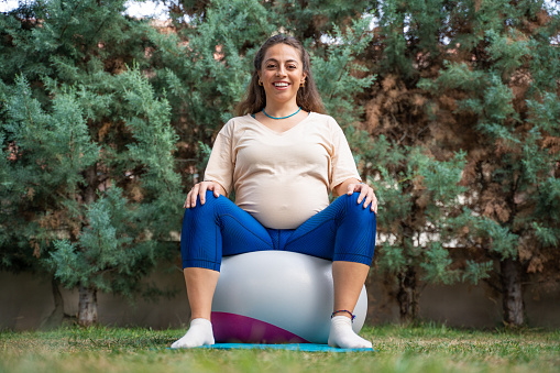 Young pregnant doing pilates outdoors is smiling on pilates ball