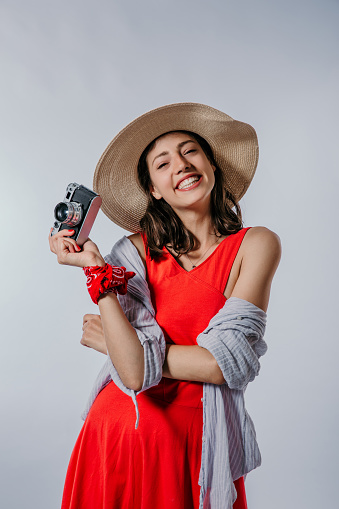 A photo of a cute young woman standing in a red dress covering with shirt, wearing a summer hat and a bandanna over her hand while holding a retro camera and looking at the camera with a big smile on her face. Isolated in a studio