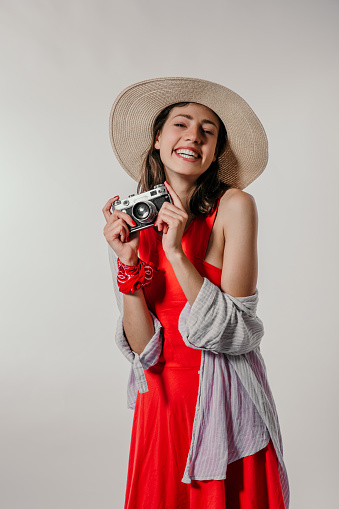 A photo of a cute young adult standing in a red dress covering with shirt, wearing a summer hat and a bandanna over her hand and holding a retro camera isolated in a studio. Radiating happiness