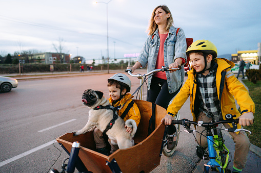 Photo of a cute little boy riding with his dog in a cargo bike, accompanied by his mother and brother, and enjoying the warm afternoon in the city.