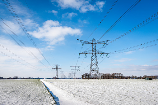 Power lines and pylons in a winter landscape. A layer of untouched snow is on the field. The sky is bright blue with some smal white clouds. The photo was taken in the Dutch province of Noord-Brabant.