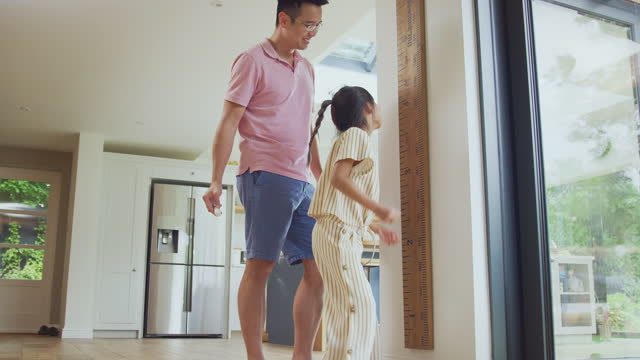 Asian Father Measuring Daughter On Wall Scale At Home As She Stands On Tip Toes