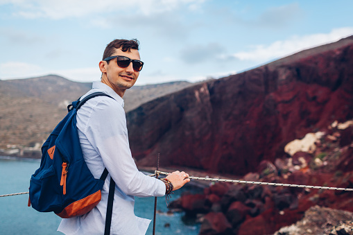 Tourist enjoying Red beach, Aegean sea and mountains landscape from view point in Akrotiri, Santorini island, Greece. Happy man backpacker traveling