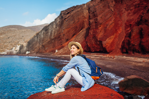 Young traveler woman relaxing sitting on Red beach rock on Santorini island Greece admiring seaside view, mountain landscape. Tourist enjoys summer vacation