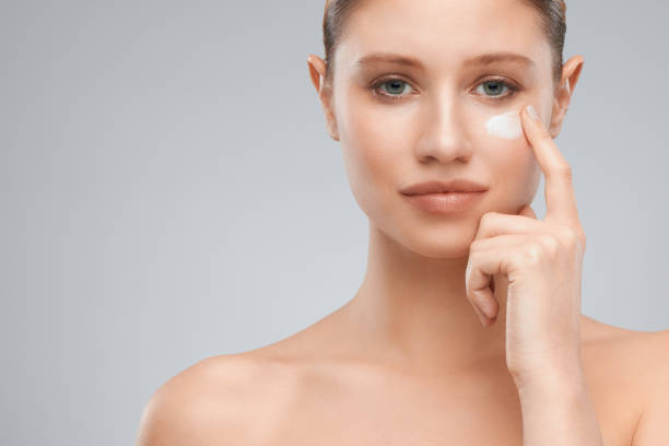 istock Beautiful blond woman putting moisturizer on her face while standing in studio. 1302695740
