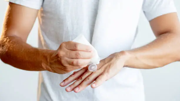 Man bodycare. Male cosmetology. Grooming routine. Unrecognizable athletic guy in white t-shirt applying hand cream from tube isolated on blur light background.