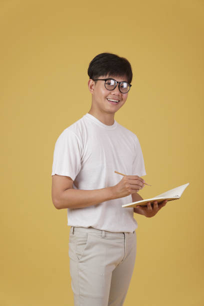 istock happy cheerful young asian man student dressed casually wearing eyeglasses writing note isolated on yellow studio background 1302708368