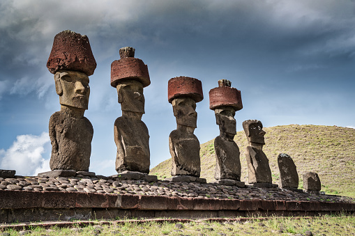 Ahu Nau Nau Ancient Moai Statues, Seven Moais side by side in a row with ancient stone headgear at Anakena Beach under moody summer sky looking towards the beach and pacific ocean. Ahu Nau Nau - Anakena - is also known as Haŋa rau o te 'ariki - The Bay of the King Beach. Anakena Beach, Rapa Nui National Park, Hanga Roa, Easter Island, Isla de Pascua, Polynesia, Oceania, Chile