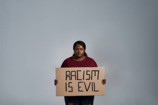 serious plus size young african american woman in casual clothes looking at camera, holding racism is evil banner in front of her, posing isolated over gray background - anti racism imagens e fotografias de stock