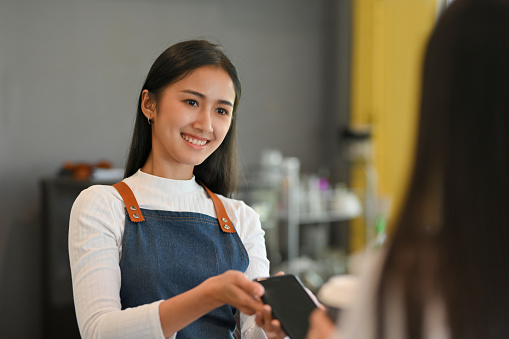 Portrait of young beautiful barista with apron holding over smartphone to help customer pay for purchase a coffee