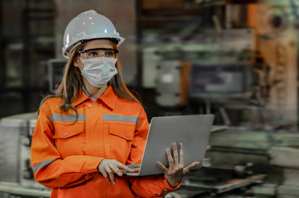 Portrait of Engineer wearing the face mask and holding laptop with blurred factory machinery background stock photo