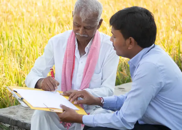 Photo of Selective focus on farmer, Banker or corporate officer getting sign from farmer while sitting near the farmland - concept of cotract farming, business deal and farm loan approval.