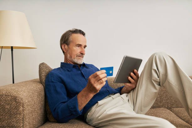 homem barbudo bonito segurando um tablet digital e cartão de crédito enquanto sentado em um sofá em casa - idoso na internet - fotografias e filmes do acervo