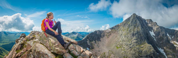 Young woman hiker looking over at the jagged north face and iconic summit of Ben Nevis, Britain’s highest mountain, from the rocky ridge of the CMD Arete, Highlands, Scotland.