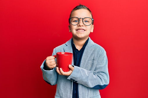 Little kid boy wearing glasses drinking from a red mug looking positive and happy standing and smiling with a confident smile showing teeth