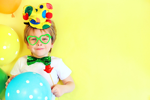 Funny smiling child clown on yellow background with multicolored balloons, copy space. April fools' day celebration concept. Birthday party concept