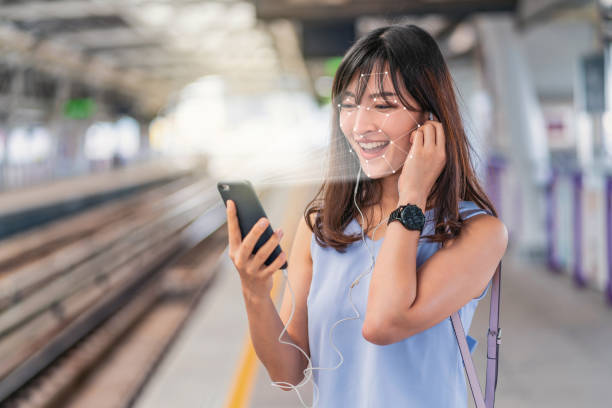 Asian woman using face recognition via smart mobile phone and listening the music at Railroad Station Platform, Biometric Verification and artificial intelligence concept stock photo