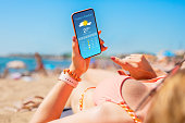 Woman sunbathing on the beach in hot summer day and checking weather forecast on mobile phone.