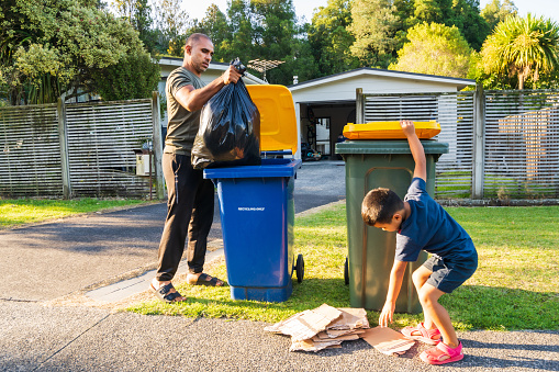 Kids helping father to sort out the rubbish by putting rubbish and recyclable material in recycle bins in front of house in Auckland, New Zealand.