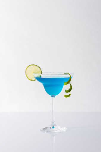 Blue alcoholic drink in cocktail glass with lime slice and lime zest ribbon isolated on white and gray background