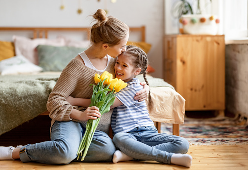 Happy woman with bouquet of tulips in hand embracing and kissing cute little daughter while receiving greetings with Mothers day in bedroom at home