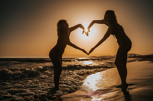 Two young female friends standing on the beach and holding hands in heart shape with beautiful summer sunset in background