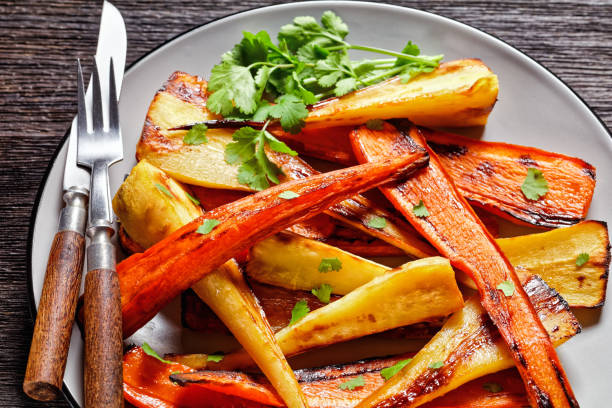 marokkanisches röstes wurzelgemüse: parsnip und karotten mit ras el hanout gewürz: zimt, kurkuma, anis serviert auf einem teller mit frischem koriander und besteck auf holzhintergrund, ansicht von oben, nahaufnahme - parsnip vegetable winter food stock-fotos und bilder