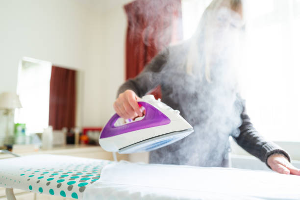 Color image depicting a senior caucasian woman in her 70s ironing shirts in her bedroom at home. Room for copy space.